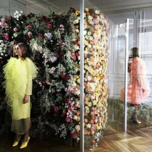 Modèle en robe jaune volée devant un mur de fleurs colorées, dans un espace élégant et lumineux.