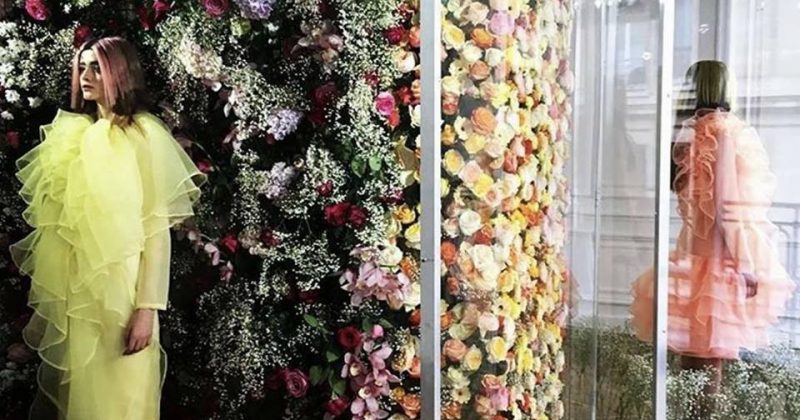 Modèle en robe jaune volée devant un mur de fleurs colorées, dans un espace élégant et lumineux.