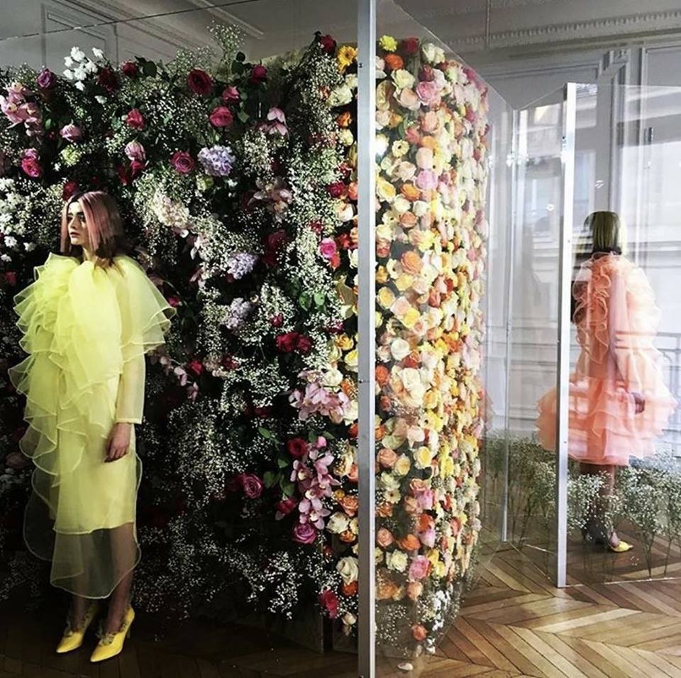 Modèle en robe jaune volée devant un mur de fleurs colorées, dans un espace élégant et lumineux.