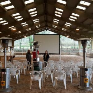 Salle équipée de chaises blanches, chauffages et écran, préparée pour un événement au Haras de la Cense.