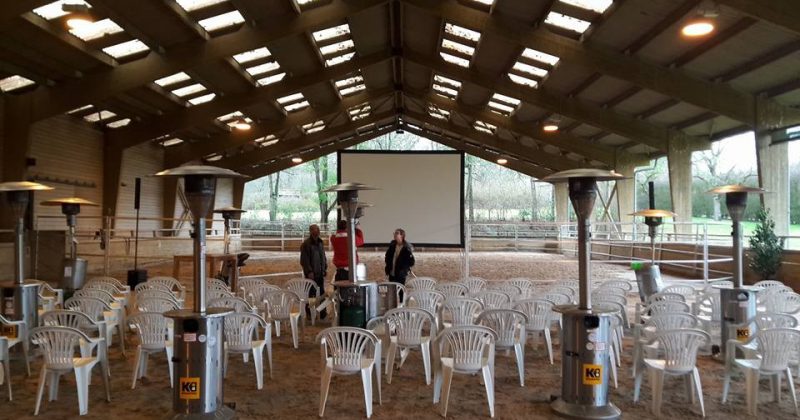 Salle équipée de chaises blanches, chauffages et écran, préparée pour un événement au Haras de la Cense.