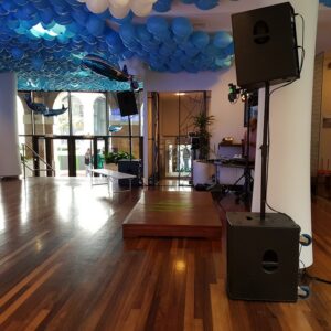 Espace événementiel avec podium, matériel de sonorisation, éclairage et décorations de ballons bleu et blanc.