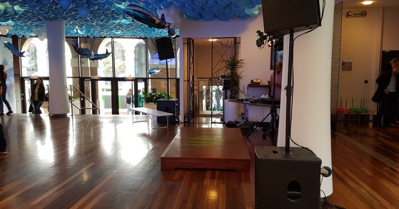 Espace événementiel avec podium, matériel de sonorisation, éclairage et décorations de ballons bleu et blanc.