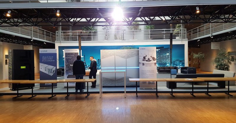 Stand d'Atos/Bull à une exposition, présentant des équipements de son et d'éclairage. Deux personnes discutent devant un panneau d'affichage.
