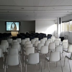 Salle de conférence avec rangées de chaises face à un écran de projection et un éclairage naturel.