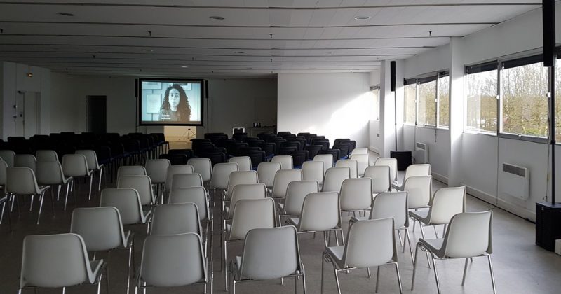 Salle de conférence avec rangées de chaises face à un écran de projection et un éclairage naturel.