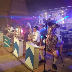Groupe de musiciens en tenue traditionnelle bavaroise jouant sur scène, avec éclairages colorés et instruments variés.