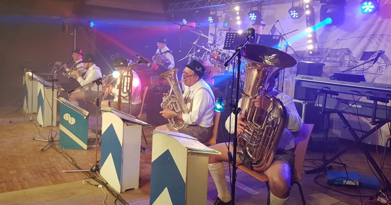 Groupe de musiciens en tenue traditionnelle bavaroise jouant sur scène, avec éclairages colorés et instruments variés.