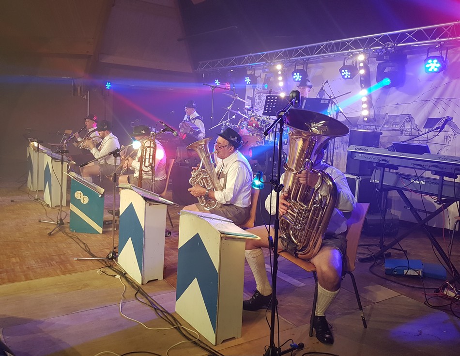 Groupe de musiciens en tenue traditionnelle bavaroise jouant sur scène, avec éclairages colorés et instruments variés.