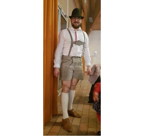 Homme en tenue traditionnelle avec chapeau, bretelles et short, posé dans un intérieur chaleureux.
