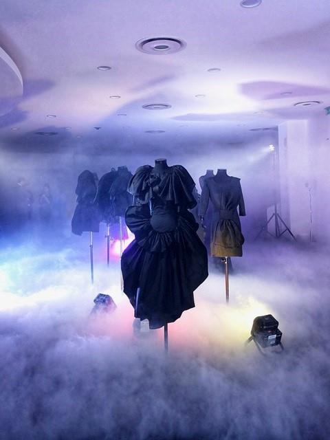 Robe noire présentée dans un espace brumeux, avec éclairage coloré et mannequins stylisés lors de l'événement mode avec Xuan.