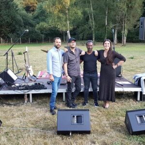 Quatre personnes posent devant une scène équipée de matériel audio et d'éclairage, pour un événement en plein air.