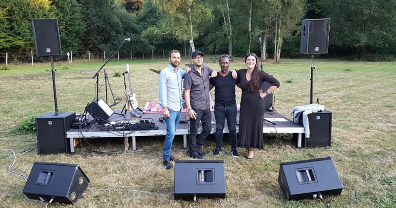 Quatre personnes posent devant une scène équipée de matériel audio et d'éclairage, pour un événement en plein air.