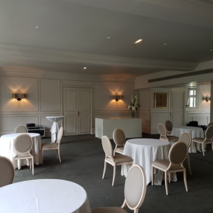 Intérieur d'une salle d'événements, avec tables rondes, chaises claires et éclairage doux.