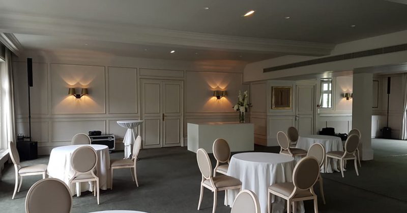 Intérieur d'une salle d'événements, avec tables rondes, chaises claires et éclairage doux.