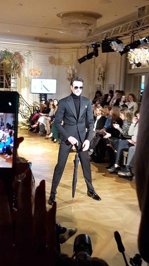 Homme en costume noir avec lunettes de soleil, posant avec un parapluie sur un podium de défilé de mode au Ritz.