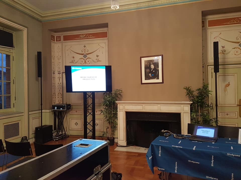 Salle de conférence avec équipement audiovisuel et présentation sur un écran, décorée et équipée pour un événement.