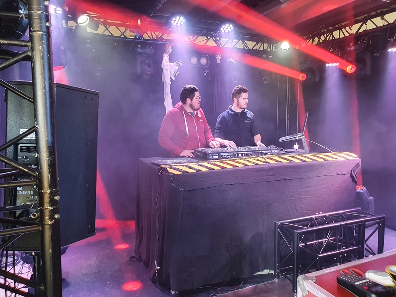 Deux DJs derrière un platine, avec effets de lumière et fumée de scène. Équipement de sonorisation en arrière-plan.