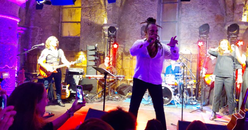 Concert de Roddy Julienne avec des musiciens sur scène, éclairage coloré et public filmant l'événement.