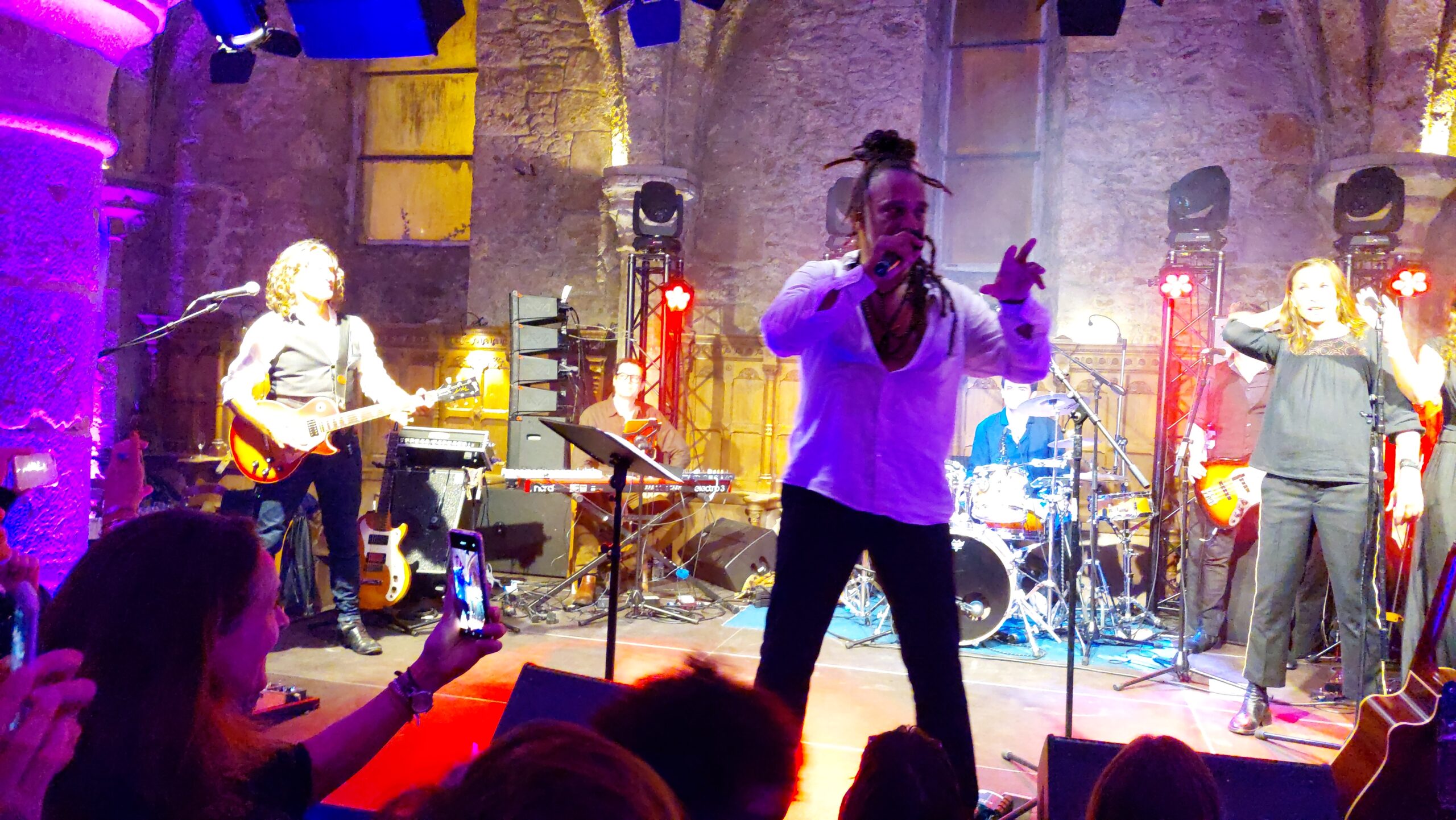 Concert de Roddy Julienne avec des musiciens sur scène, éclairage coloré et public filmant l'événement.
