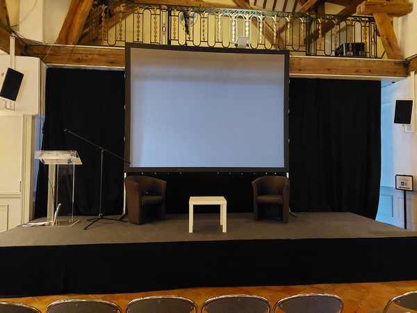 Scène avec un écran, deux fauteuils et une table, prête pour un événement à Grand Maison le 17 mai 2022.