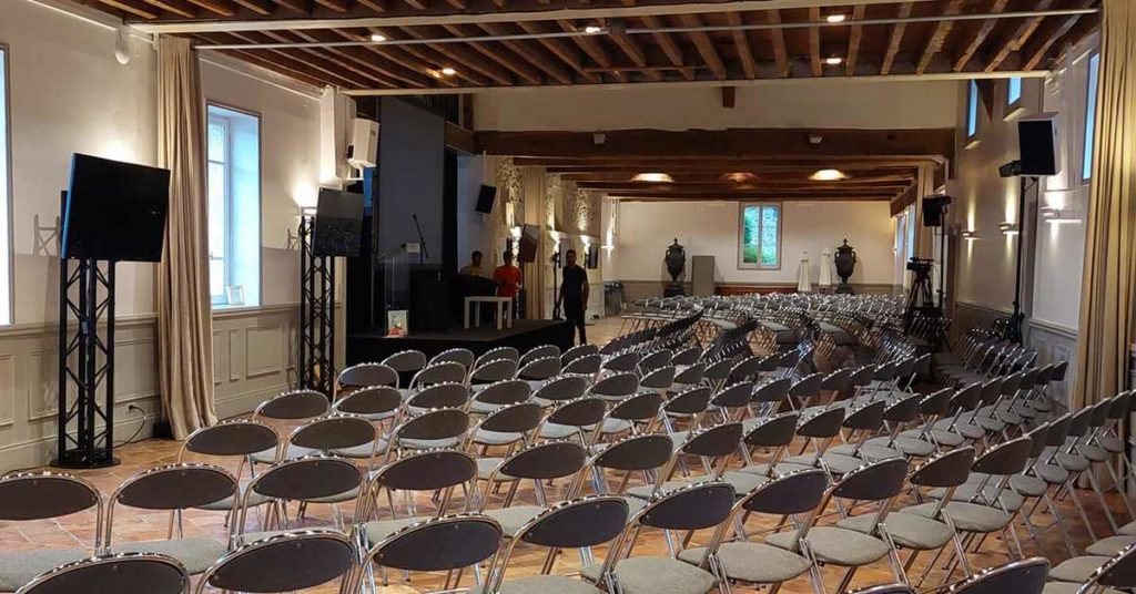 Salle de conférence avec rangées de chaises, écrans et scène, préparée pour un événement le 17 mai 2022 par Symbiose Organisation.