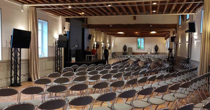 Salle de conférence avec rangées de chaises, écrans et scène, préparée pour un événement le 17 mai 2022 par Symbiose Organisation.