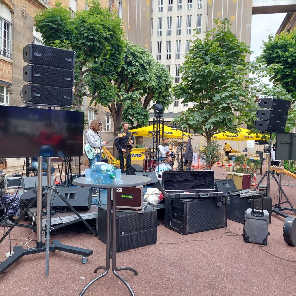 Installation de matériel de sono pour un concert en extérieur, avec musiciens sur scène et équipements audio visibles.