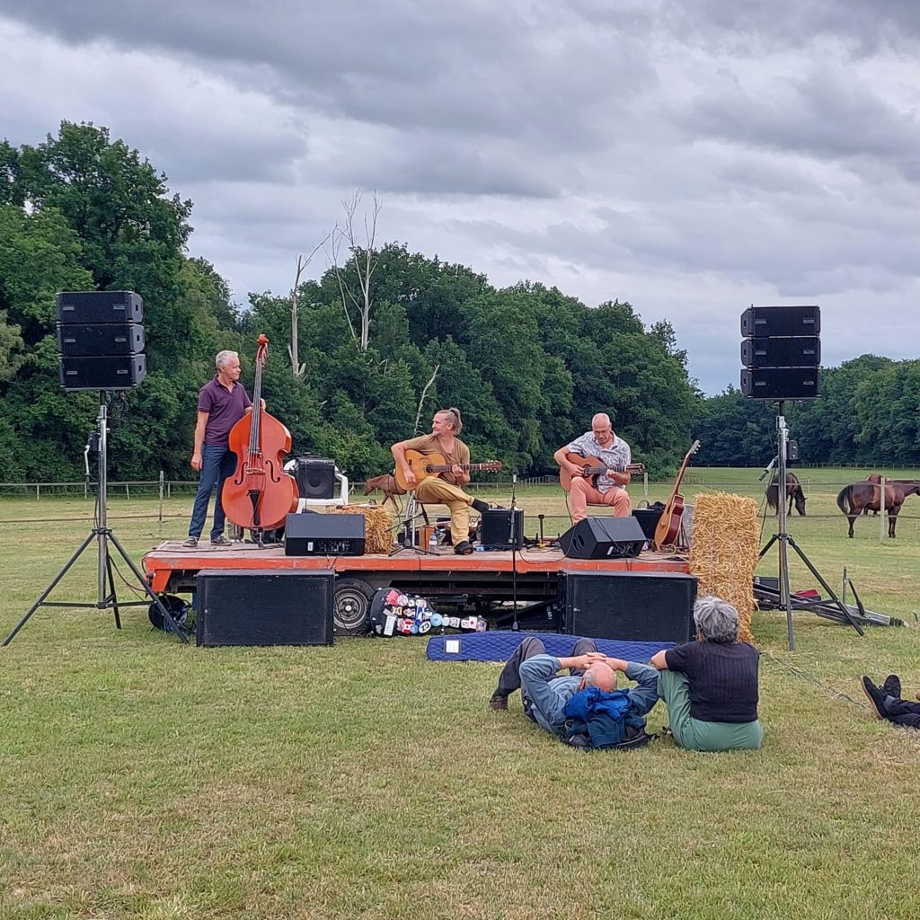 Concert de jazz en plein air avec trois musiciens sur scène, spectateurs se détendant sur l'herbe, ambiance paisible.