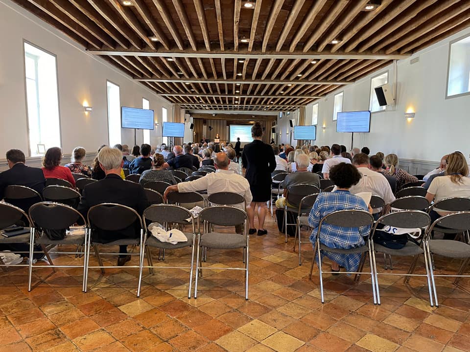 Salle avec des chaises disposées, un public attentif et deux écrans affichant des présentations lors d'un événement organisé par Symbiose.