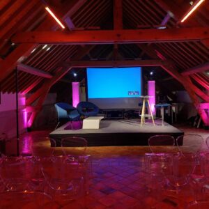 Scène prête pour un événement au Grand Maison, avec éclairage violet, chaises transparentes et équipement audiovisuel.
