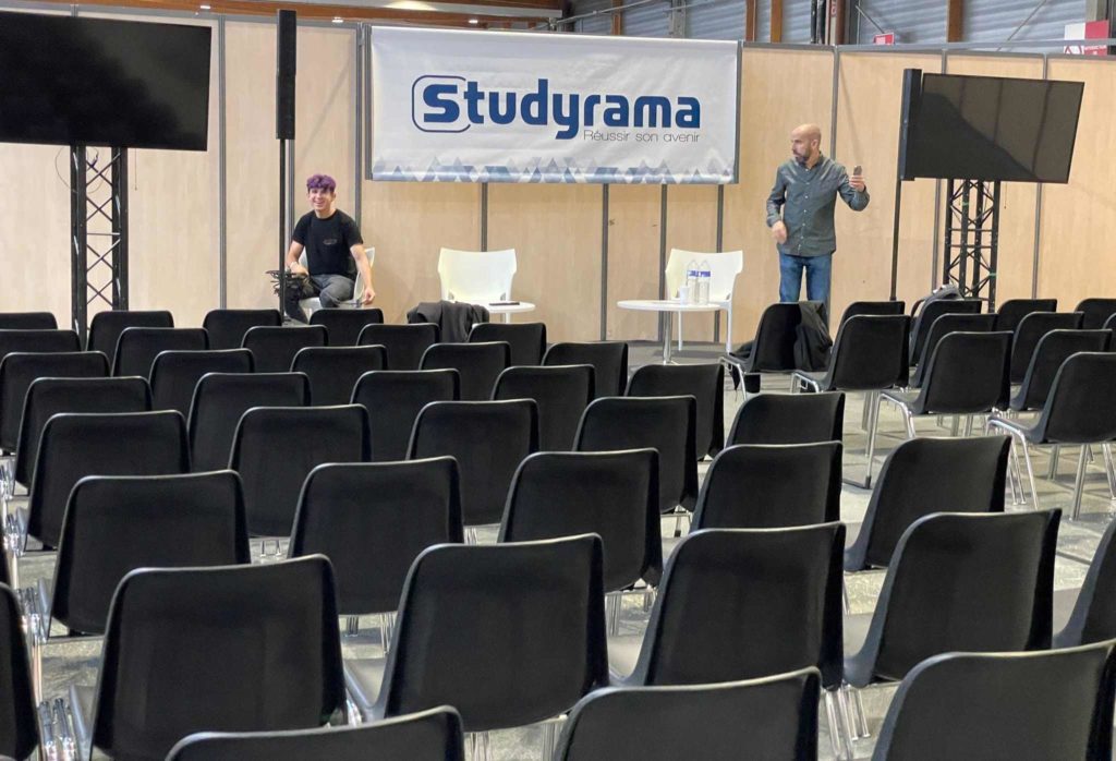 Deux personnes s'affairent dans un salon d'expo Studyrama, avec des chaises noires et des écrans en arrière-plan.