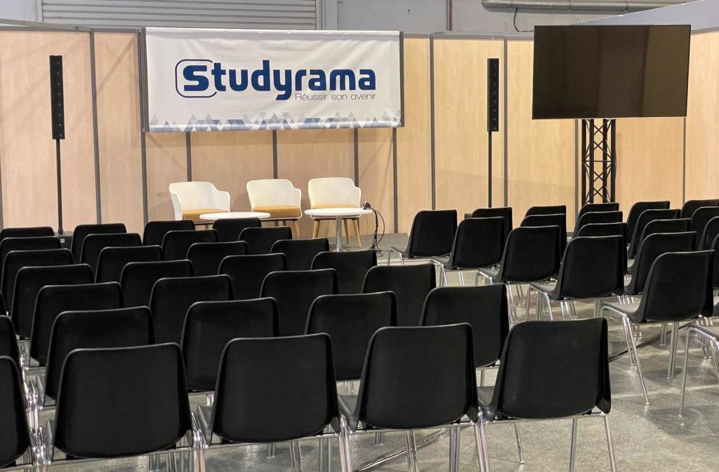 Salle d'exposition avec des chaises noires, panneau Studyrama et espace pour une présentation, lors d'un événement à Paris.