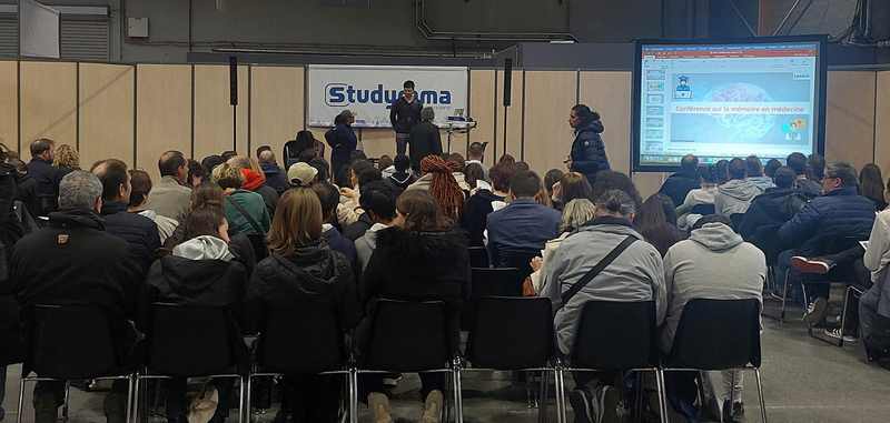 Audience réunie lors d'une présentation au salon d'exposition à Paris, avec intervenant et écran de projection.