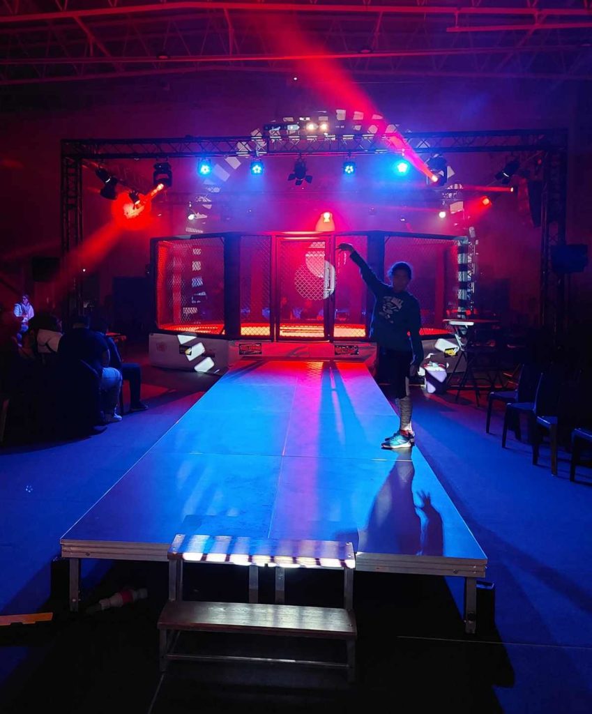 Scène illuminée pour une compétition de MMA, avec une cage au centre et un technicien vérifiant les équipements.