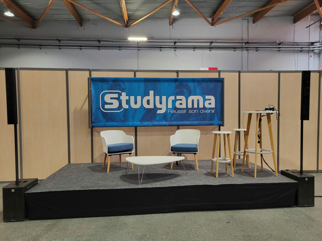 Scène avec logo Studyrama, deux chaises, tables et équipement de sonorisation dans un salon d'expo.
