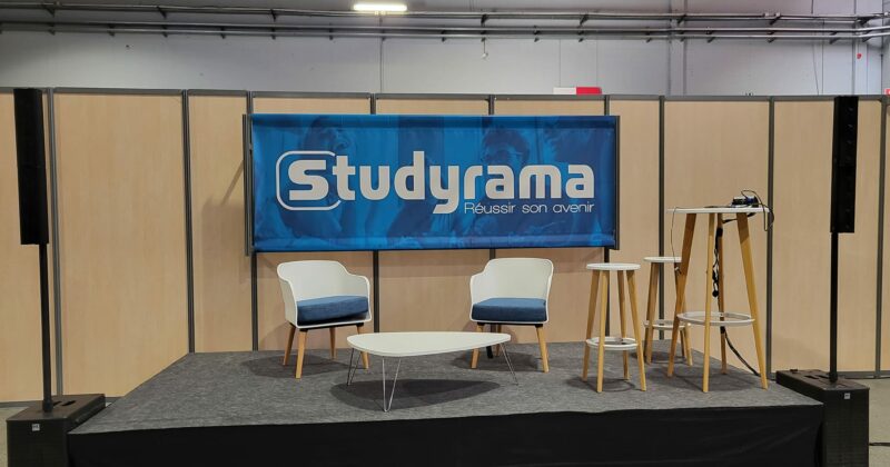 Scène avec logo Studyrama, deux chaises, tables et équipement de sonorisation dans un salon d'expo.