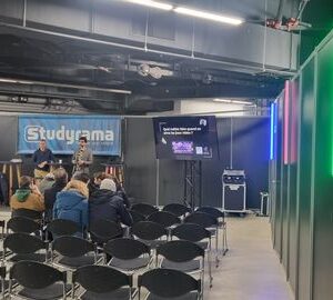 Salon Studyrama 2024 avec une salle de conférence, des spectateurs et un écran de présentation. Éclairage coloré en arrière-plan.