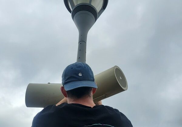 Technicien installant un haut-parleur dans un lampadaire, portant un t-shirt de Symbiose Organisation.