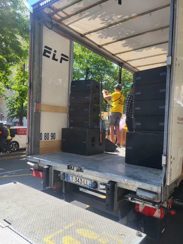 Char N°2 de la marche de Jésus, équipé d'un système de sonorisation sur un camion, avec des participants en arrière-plan.