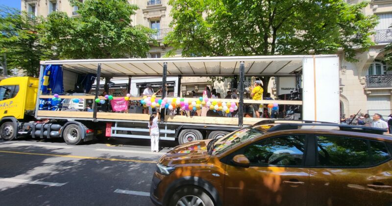 Char décoré de ballons pour la marche de Jésus, bien sonorisé, transporté par un camion jaune. Activité festive sur la route.