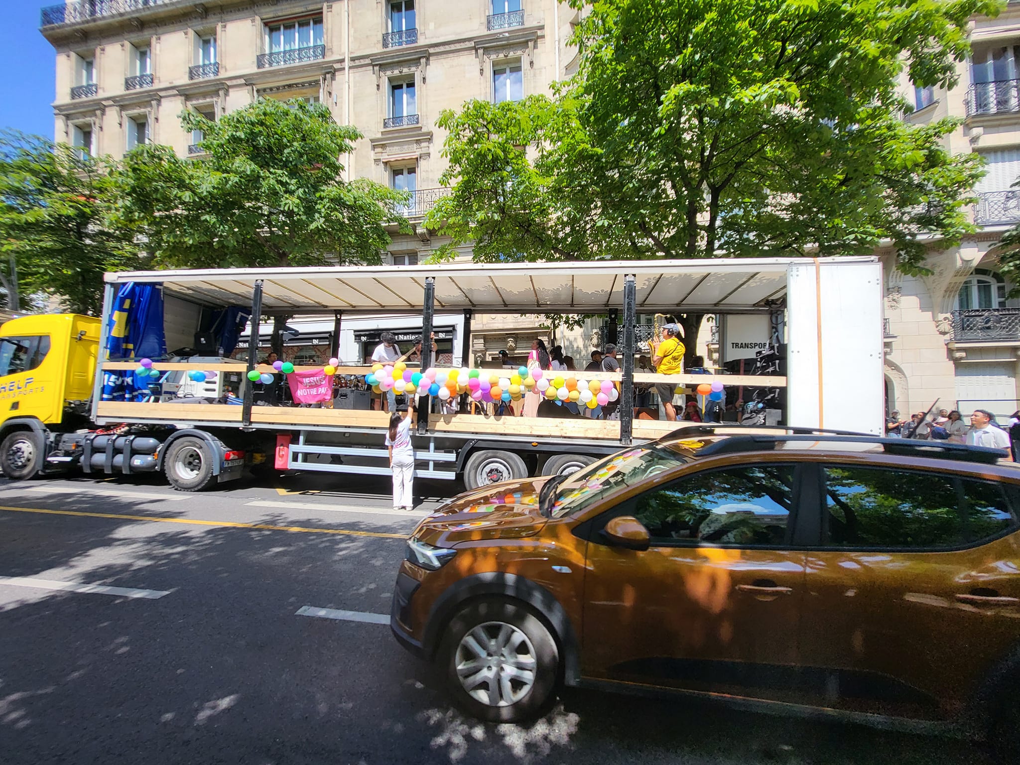Char décoré de ballons pour la marche de Jésus, bien sonorisé, transporté par un camion jaune. Activité festive sur la route.