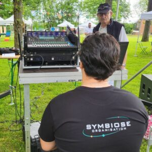 Technicien de son gérant un matériel audio pour les Olympiades de Neauphle-le-Château, avec une vue sur un parc et des tentes.
