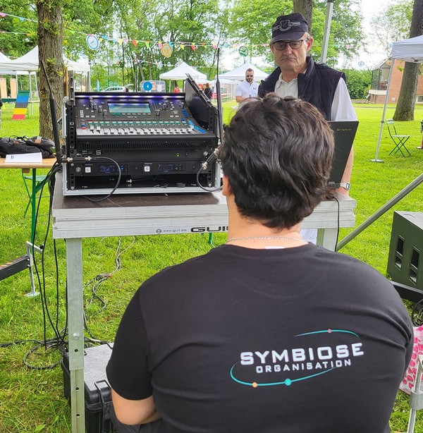 Technicien de son gérant un matériel audio pour les Olympiades de Neauphle-le-Château, avec une vue sur un parc et des tentes.