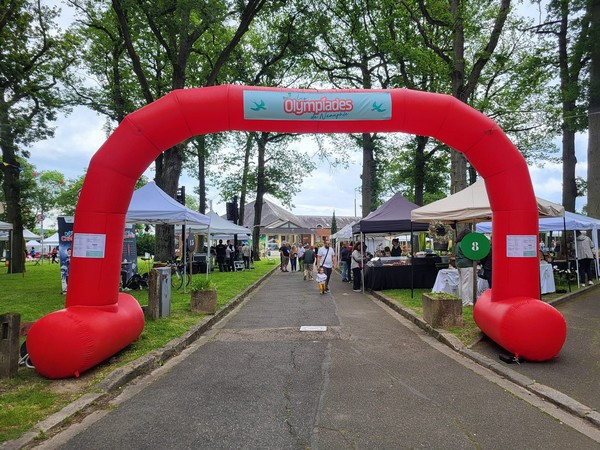 Arche rouge marquant l'entrée des Olympiades de Neauphle-le-Château le 26 mai 2024, entourée de stands et d'un parc.
