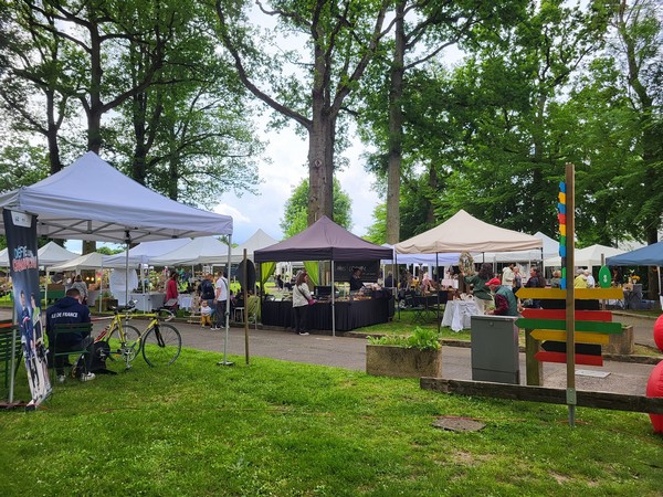 Des stands de marché sous des tentes dans un parc, avec des visiteurs et des arbres environnants. Événement à Neauphle-le-Château.