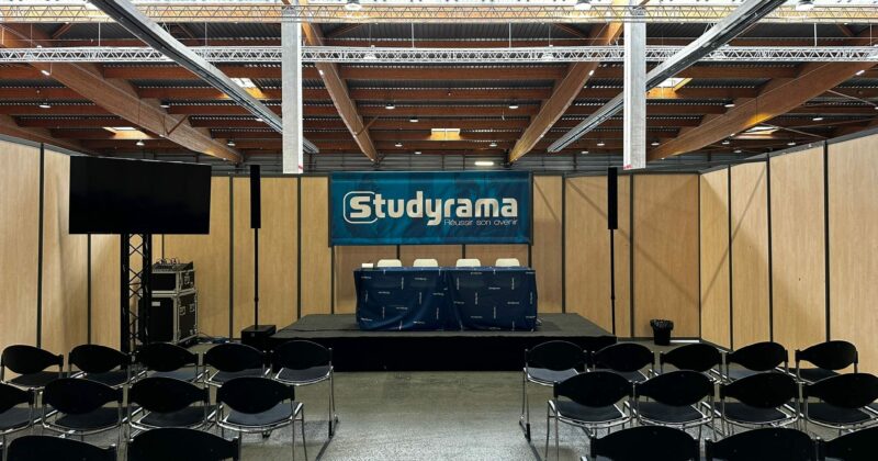 Stand Studyrama avec des chaises noires, un podium et un écran, prêt pour un événement au sein d'un espace ouvert.
