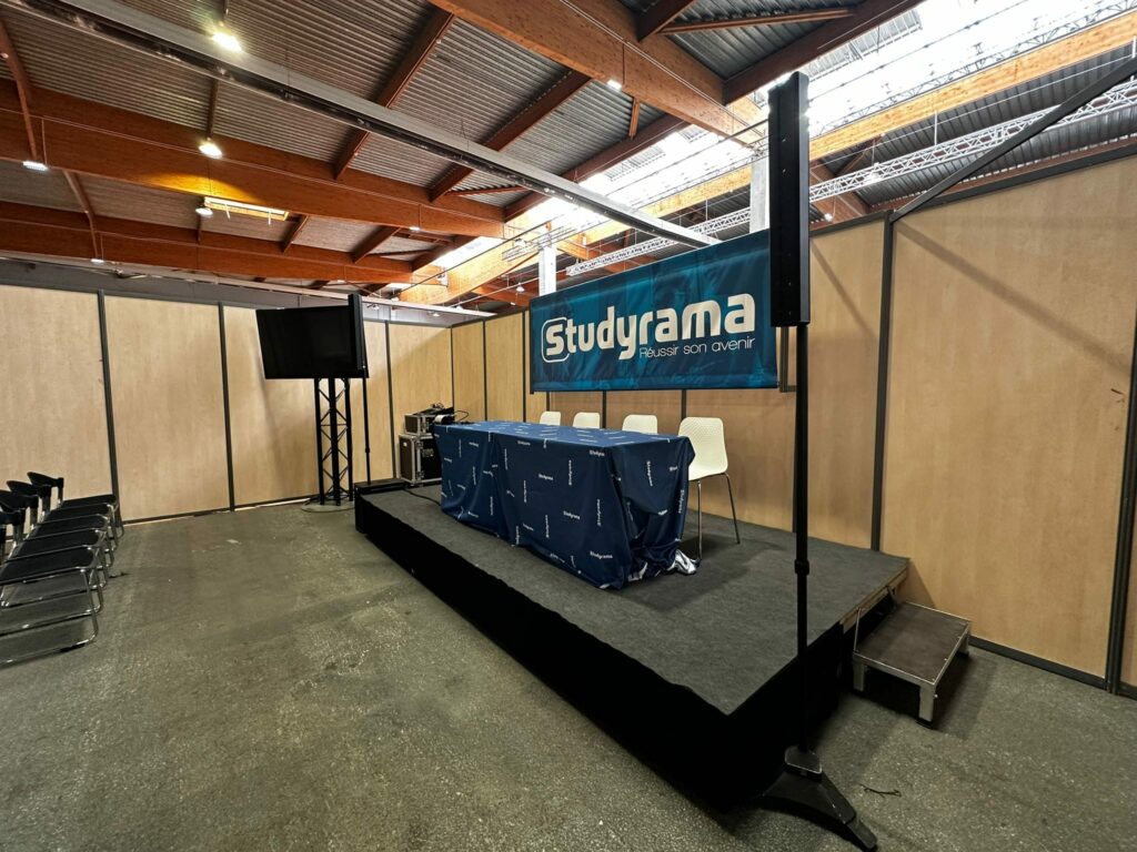 Stand d'exposition pour Studyrama avec une scène, des chaises, un écran et des équipements de sonorisation.