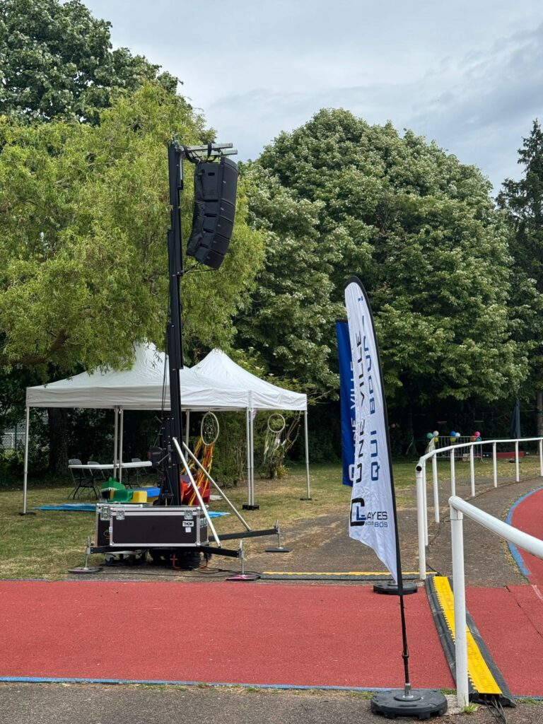 Équipement de sonorisation monté sur un mât, avec une tente et un drapeau d'entreprise, en arrière-plan d'un espace extérieur.