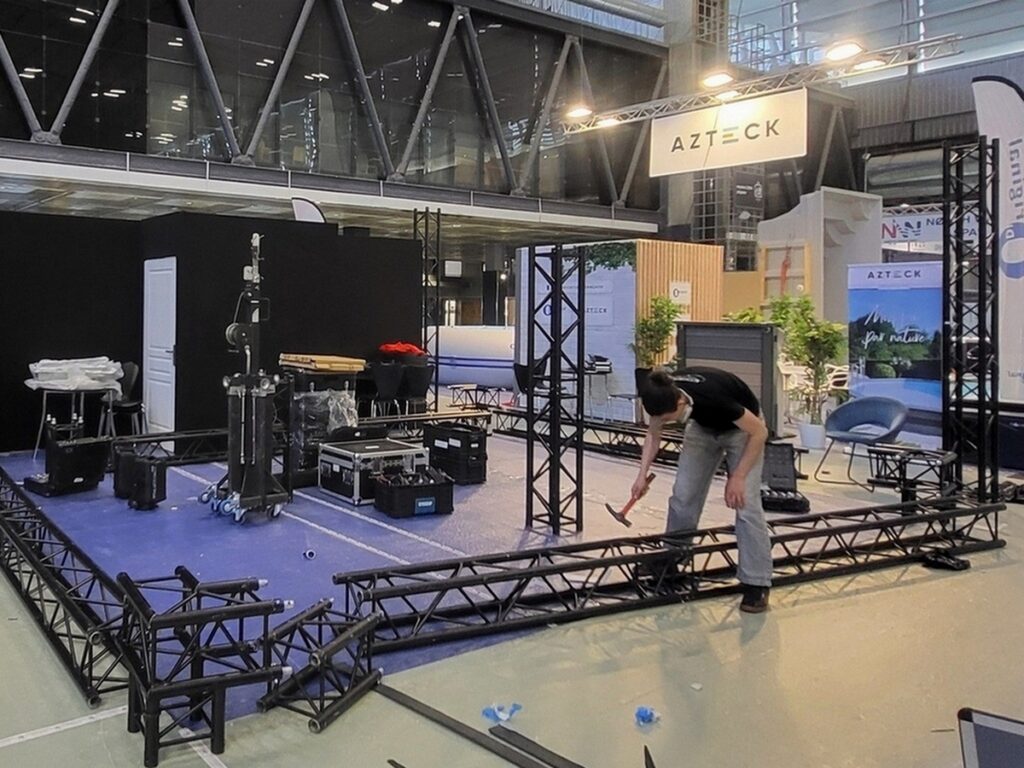 Montage de matériel sonore et d'éclairage par un technicien dans un espace événementiel, avec des structures et des équipements visibles.